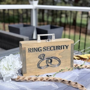 May include: A wooden ring security box with a silver handle and clasp. The box is engraved with the words "RING SECURITY" and features a diamond ring design. The box is sitting on a white surface.