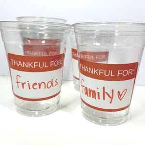May include: Clear plastic cups with orange and white labels. The labels read "THANKFUL FOR: friends" and "THANKFUL FOR: family". The word "family" has a red heart symbol. The cups are for a party or gathering.