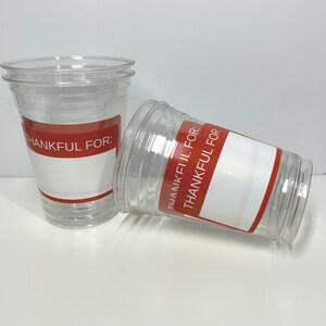 May include: Two clear plastic cups with red and white labels. The labels read "THANKFUL FOR:" and have space for writing. One cup is upright, the other is tilted on its side.