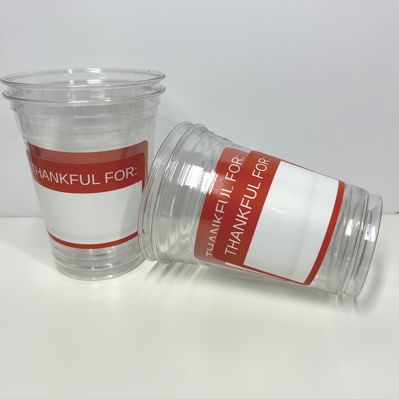 May include: Two clear plastic cups with red and white labels. The labels read "THANKFUL FOR:" and have space for writing. One cup is upright, the other is tilted on its side.