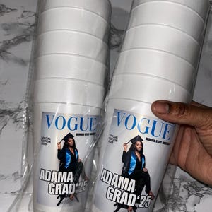 May include: Two stacks of white foam cups, each with a custom label. The label features the Vogue logo, a photo of a graduate, and the text "ADAMA GRAD '25". The cups are packaged in clear plastic.