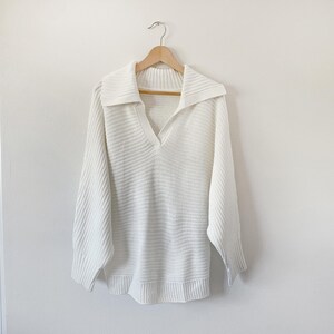 May include: A white, long-sleeved sweater with a wide, ribbed collar and a V-neck. The sweater is hanging on a wooden hanger against a plain white background. The knitwear has a textured appearance.