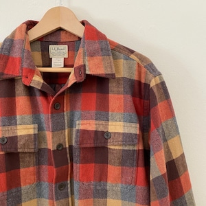 May include: A button-down flannel shirt in a traditional fit, featuring a classic plaid pattern in shades of red, orange, brown, and tan. The shirt has two chest pockets and a collar. The label reads "LLBean".