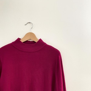 May include: A magenta-colored turtleneck sweater hangs on a wooden hanger against a white background. The sweater has a ribbed collar and long sleeves. The material appears soft and comfortable.