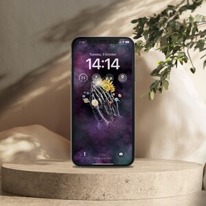 Pode incluir: Um smartphone com fundo preto e um gráfico de duas mãos juntas com um sol e planetas ao redor. A hora é 14:14 e a data é terça-feira, 3 de outubro. O telefone está em uma superfície marrom claro.