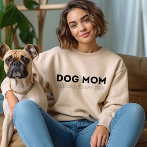 Può includere: Una donna indossa una felpa beige con la scritta "DOG MOM ALL DAY. EVERY DAY."  È seduta su un divano con un piccolo cane marrone chiaro.