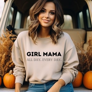 Könnte beinhalten: Ein beiger Pullover mit dem Aufdruck "GIRL MAMA ALL DAY. EVERY DAY." in Schwarz. Das Model trägt eine blaue Jeans und sitzt auf der Ladefläche eines Lastwagens.