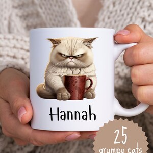 Może przedstawiać: Biały kubek ceramiczny z ilustracją zrzędliwego kota trzymającego czerwony kubek. Kubek jest spersonalizowany imieniem "Hannah" i ma brązową naklejkę z tekstem "25 grumpy cats".