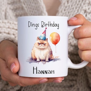 Może przedstawiać: Biały kubek ceramiczny z obrazkiem zrzędliwego kota w czapce urodzinowej trzymającego balon. Na kubku nadrukowany jest tekst "Dings Birthday" i "Hannah".