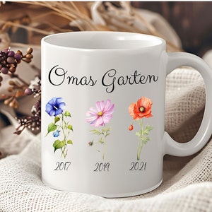 May include: White ceramic mug with a black inscription "Omas Garten" and three watercolor flowers with the years 2017, 2019, and 2021 printed below them.
