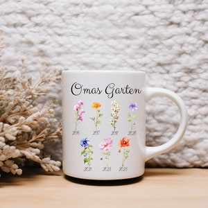 May include: White ceramic mug with the text "Omas Garten" and a floral design. The flowers are arranged in rows with the years 2015, 2017, 2018, 2020, 2021, 2023, and 2023 printed below them.