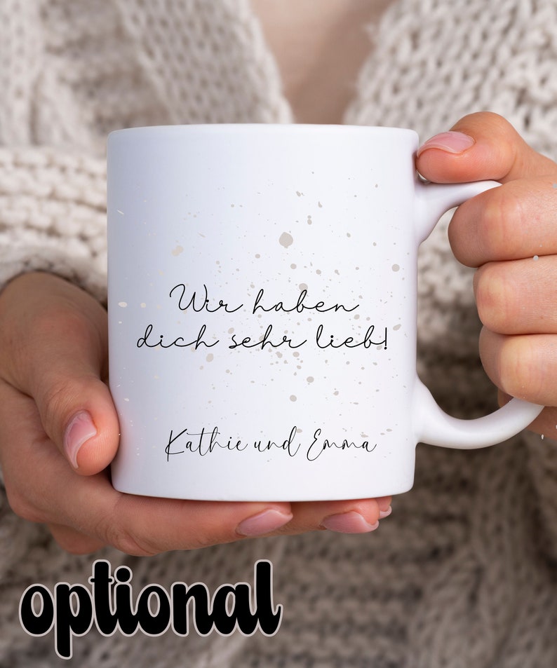 May include: White ceramic mug with a black and white splatter design. The mug has the text "Wir haben dich sehr lieb!" and "Kathie und Emma" printed on it.