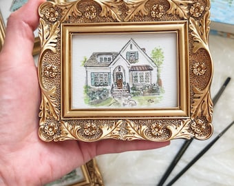 May include: A small watercolor painting of a white house with a brown roof and green shutters, housed in an ornate, rectangular gold-colored frame with floral and leaf relief details. The artwork features a front door, windows, and landscaping.