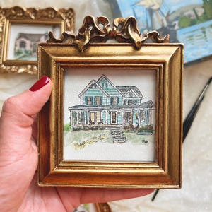 May include: A small square watercolor painting of a two-story house with teal siding and a dark roof, set inside an ornate gold frame with a decorative bow at the top. The house features a front porch, multiple windows, and a stone walkway.