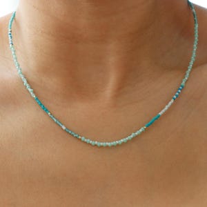 May include: Delicate beaded necklace featuring a mix of light blue, turquoise, and white beads. The necklace is worn against a dark background, highlighting the colors of the beads. A simple and elegant piece of jewelry.