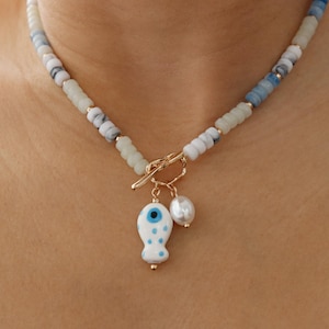 May include: A necklace with a gold clasp and a charm. The necklace has white, blue, and green beads. A gold charm with a white and blue evil eye design and a white pearl hangs from the clasp.