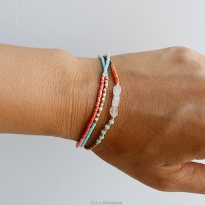 May include: A multi-strand bracelet featuring small seed beads in shades of coral, light blue, white, and brown, accented with three larger, translucent, pale pink oval-shaped beads. The bracelet is composed of several thin, delicate strands.