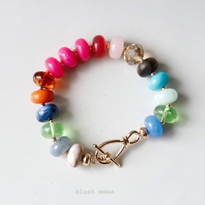 May include: A colorful beaded bracelet with a gold-tone clasp. The bracelet features a variety of beads in shades of pink, red, orange, blue, green, and clear. The clasp is a toggle style. The text "blush emma" is at the bottom.