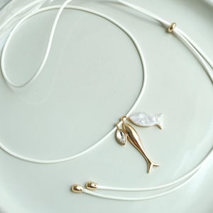 May include: A white cord necklace with gold and pearl accents. The necklace features a gold fish pendant, a pearl fish pendant, and a clear crystal pendant. The necklace is displayed on a white plate.