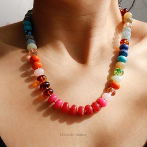 May include: A colorful beaded necklace with a gradient of colors, from red to blue, is displayed. The necklace features a variety of bead shapes and sizes, including round and oval beads. The necklace is worn against a person's skin, with a black top visible.