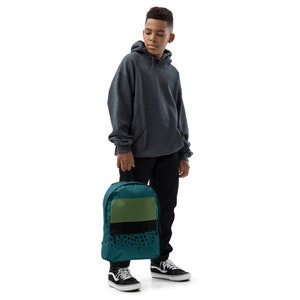 May include: Teal backpack with a green and black design. The backpack has a rectangular green section at the top, a black stripe with black dots, and a teal base. The backpack is held by a person wearing a gray hoodie and black pants.