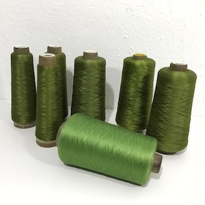 May include: Seven spools of olive green thread are arranged on a white surface. The spools are cylindrical, with a cardboard core and tightly wound thread. One spool lies horizontally in the foreground, while the others stand upright.