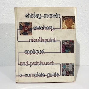 May include: A hardcover book titled Stitchery, Needlepoint, Appliqué, and Patchwork: A Complete Guide by Shirley Marein. The off-white cover features a continuous dark line connecting text and three square images of textile art projects.
