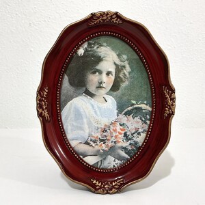 May include: An oval picture frame with a deep red finish and gold-toned ornate detailing along the edges. The frame contains a vintage-style print of a young person with wavy hair wearing a light-colored top and holding a basket of flowers.