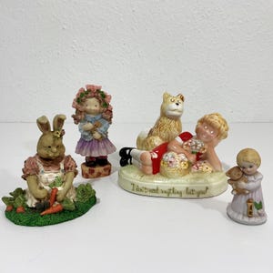 May include: A collection of four ceramic figurines: a rabbit in a garden, a girl with a floral crown, a child lying with a dog and a basket of flowers inscribed with "I don't need anything but you!", and a small child holding a teddy bear.