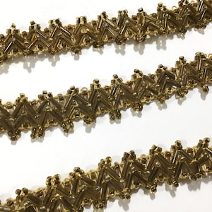 May include: Close-up of three rows of decorative trim. The trim features a zigzag pattern created with small, rectangular, golden-brown beads. The beads are sewn onto a thin, gold-colored backing, creating a textured and ornate design.