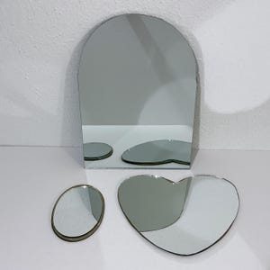 May include: A set of three mirrors consisting of one large arched mirror with a flat base, one small oval mirror, and one medium heart-shaped mirror. Each mirror features a reflective glass surface with a thin, dark-toned edge finish.