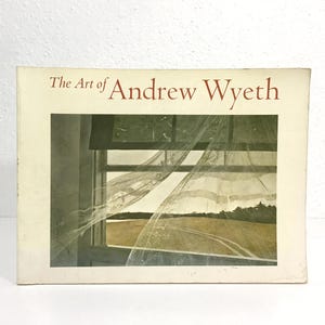May include: A book titled "The Art of Andrew Wyeth" with a painting of a window scene. The painting depicts a window with sheer curtains blowing in the wind, overlooking a field and trees. The book has a cream-colored cover.