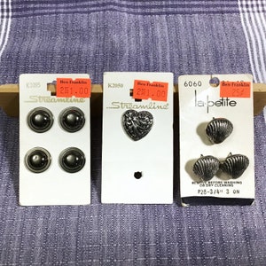 May include: Three cards with different buttons and embellishments. The first card is labeled "K2095" and features four silver buttons. The second card is labeled "K2050" and features a silver heart-shaped button with a floral design. The third card is labeled "6060" and features two silver shell-shaped buttons. The text "la petite" is printed on the card.