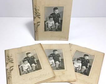 May include: Vintage greeting cards featuring black and white photographs of two young children. The cards have a sepia tone and the word "Greetings" is printed on the front. Floral illustrations adorn the left side of the cards.