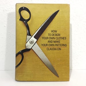 May include: A vintage book cover with a gold background and black scissors. The scissors are positioned diagonally across the cover. The book's title reads "How to Design Your Own Clothes and Make Your Own Patterns" by Claudia Ein.