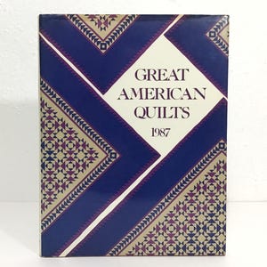 May include: A book titled "Great American Quilts 1987" with a geometric design. The cover features a white diamond shape with the title in a serif font. The background is a dark blue with a repeating pattern in beige, purple, and blue.