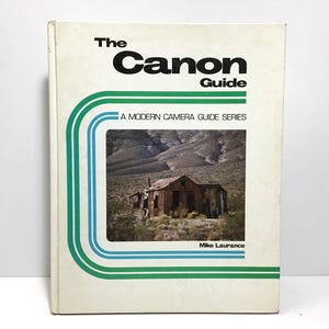 May include: A vintage book titled "The Canon Guide" with a landscape photo of a weathered building. The cover has green and blue stripes and the text "A Modern Camera Guide Series". The author's name, Mike Laurance, is at the bottom.