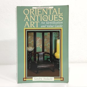 May include: A book titled "ORIENTAL ANTIQUES & ART" with the subtitle "An Identification and Value Guide." The cover is green with a gold border and features an image of antique furniture and decor. The author's name, Sandra Andacht, is at the bottom.