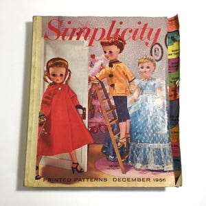 May include: Vintage Simplicity pattern book cover from December 1956. The cover features three dolls dressed in various outfits. The doll on the left wears a red coat, while the center doll is on a ladder. The cover has the word "Simplicity" in red.