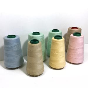 May include: A collection of colorful thread spools in various pastel shades, including blue, green, yellow, and pink. Each spool has a green base. The spools are arranged on a white surface.
