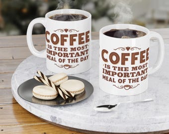 May include: Two white coffee mugs with the text "COFFEE IS THE MOST IMPORTANT MEAL OF THE DAY" in brown. The mugs are filled with coffee and are on a marble tray with a plate of macarons and a spoon.