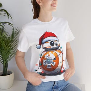 Peut inclure: T-shirt blanc avec une image de dessin anim&eacute; d'un dro&iuml;de portant un chapeau de P&egrave;re No&euml;l et entour&eacute; de lumi&egrave;res de No&euml;l. Le dro&iuml;de est blanc, orange et gris.