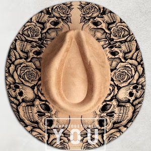 May include: Tan fedora hat with a decorative black skull and rose pattern around the brim. The hat has a teardrop crown and a wide brim. The text "UNAPOLOGETIC YOU" is printed on the brim.