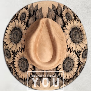 Op de afbeelding: Beige fedora hoed met een zonnebloemmotief rond de rand en een beige druppelvormige kroon. De hoed heeft de tekst "UNAPOLOGETICALLY YOU" in het wit.