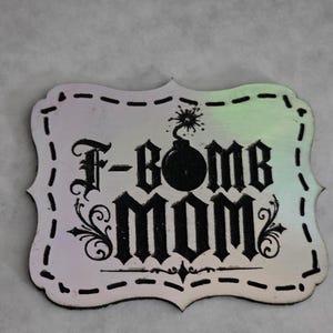May include: A rectangular iridescent plaque with rounded, scalloped edges featuring a black graphic of a bomb and the text F-BOMB MOM in a gothic font. The design includes decorative flourishes and a dashed border line around the perimeter.