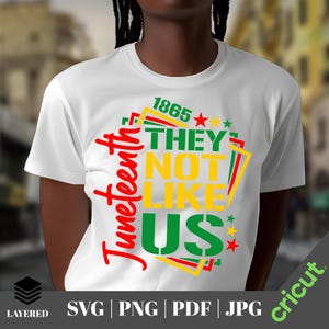 May include: White t-shirt with the text "Juneteenth" in red script. The shirt features the phrase "THEY NOT LIKE US" in green and yellow, with the year "1865" above. The design incorporates red, yellow, and green accents, and stars.