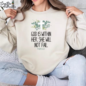 May include: A cream-colored sweatshirt with a white cross and green foliage design. The text "GOD IS WITHIN HER. SHE WILL NOT FAIL. PSALMS 46:5" is printed on the front.