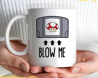 May include: White ceramic mug with a black and gray Nintendo 64 cartridge graphic and the text "BLOW ME" in black. The cartridge graphic features a red and white mushroom.