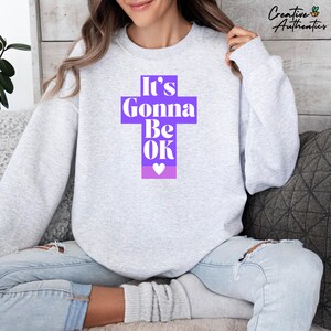 Puede incluir: Una sudadera gris claro con el texto "It's Gonna Be OK" en un diseño de cruz púrpura. El diseño incluye un pequeño corazón blanco en la parte inferior. La sudadera es de estilo casual.