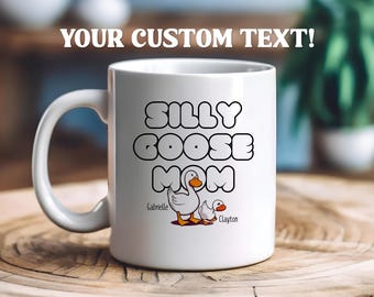 Peut inclure: Une tasse à café en céramique blanche avec un contour noir du texte "Silly Goose Mom" et deux oies de dessin animé. Les textes "Gabrielle" et "Clayton" sont écrits sous les oies.